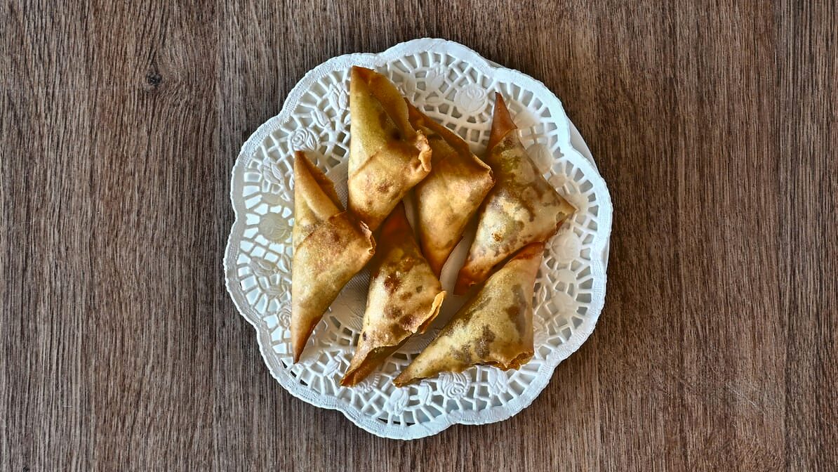 Six golden deep-fried triangular beef samosas fanned on an openwork white plate on a wood table.