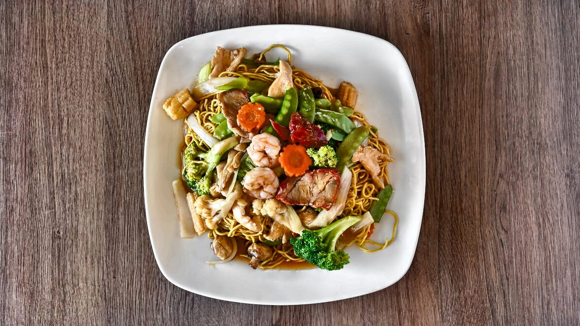 Overhead view of chow mein noodles topped with shrimp, sliced chicken, barbecue pork, broccoli, snow peas, carrots and other vegetables.