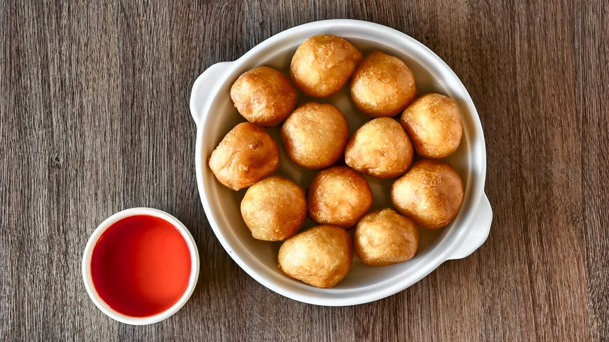 Twelve round golden battered chicken balls filling a white casserole, beside a cup of bright red sweet-and-sour sauce.