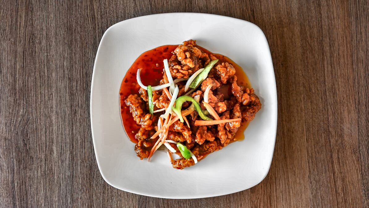 Szechuan beef from above: crisp battered beef in a glossy red-orange sauce topped with onion, carrot and green pepper strips on a square white plate.
