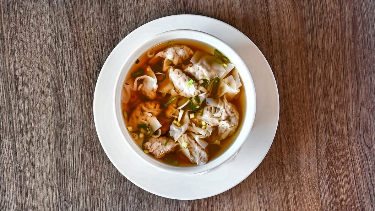 Wonton soup seen from above: folded wontons and sliced green onion in clear amber broth, in a white bowl on a white underplate on a wood table.