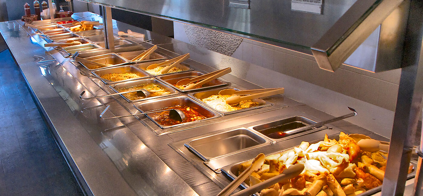 The hot buffet line seen down its length under a glass sneeze guard: steam trays with serving tongs receding into the distance, and a tray of deep-fried spring rolls and battered pieces at the near end.
