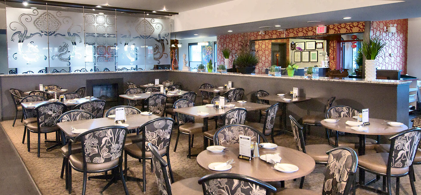 The dining room: wood-topped tables set with white plates and rolled napkins, upholstered black metal-frame chairs, etched-glass dragon panels, a red-and-gold damask wall, and framed certificates behind the granite service counter.