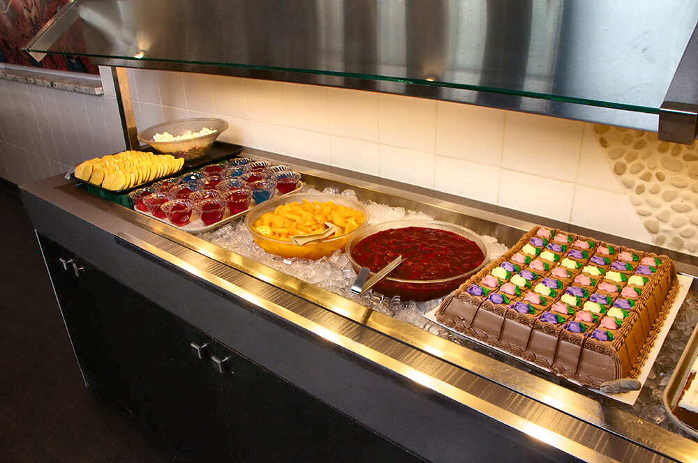 The chilled dessert bar: fanned pale-yellow rounds, rows of red and blue jelly cups, sliced peaches, cherry compote and a chocolate sheet cake with pastel icing rosettes.
