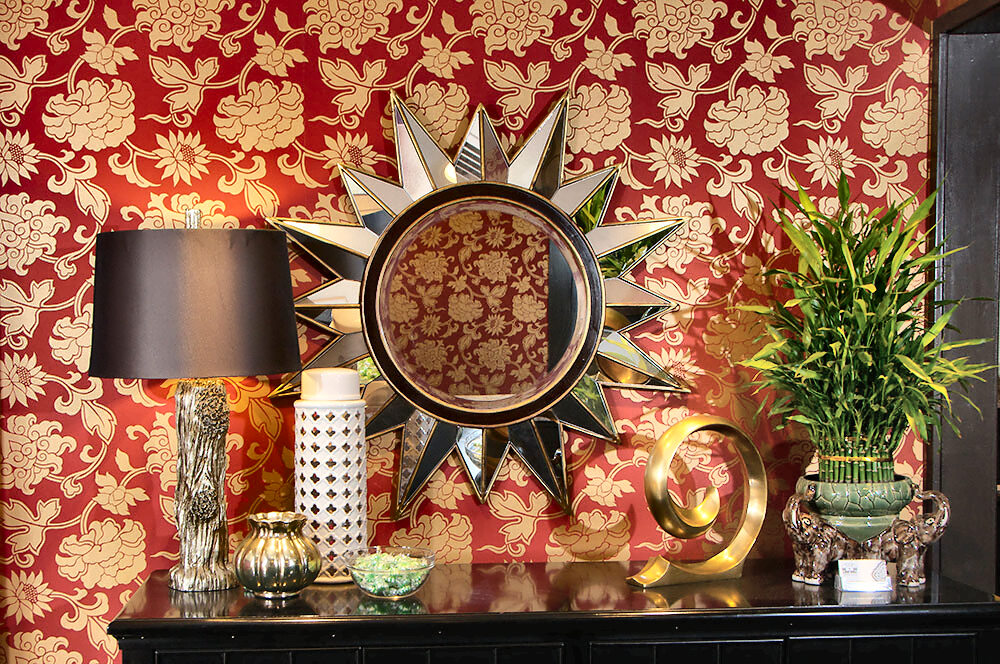 A corner of the dining room against red-and-gold floral damask wallpaper: a sunburst mirror reflecting the pattern, a brown-shaded lamp, a pierced white ceramic jar, a brass spiral and lucky bamboo in a green elephant planter.